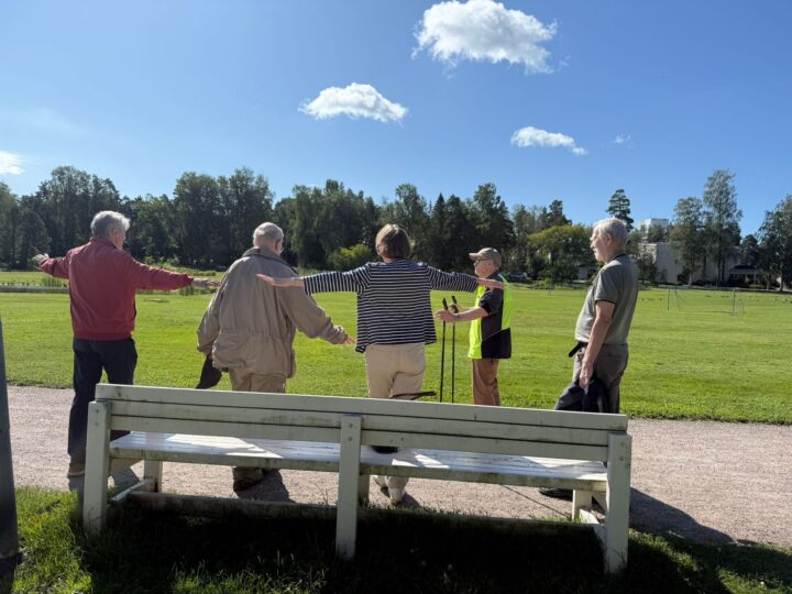 Kuvassa viisi ihmistä tekee tasapainoharjoituksia aurinkoisessa säässä nurmikentällä.