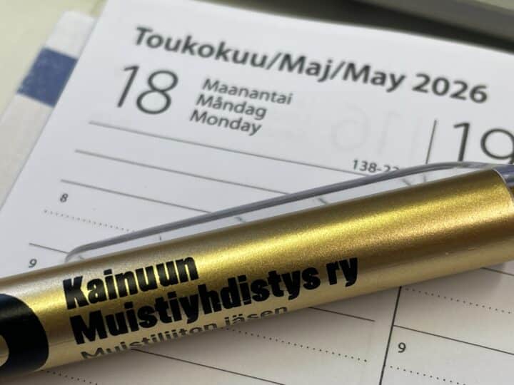 Valokuva kalenterisivulta, jossa on maanantai 18. toukokuuta 2026, päivämäärillä suomeksi, ruotsiksi ja englanniksi. Kultainen kynä, jossa on musta teksti, peittää osittain kalenteria, korostaen päivämäärää.