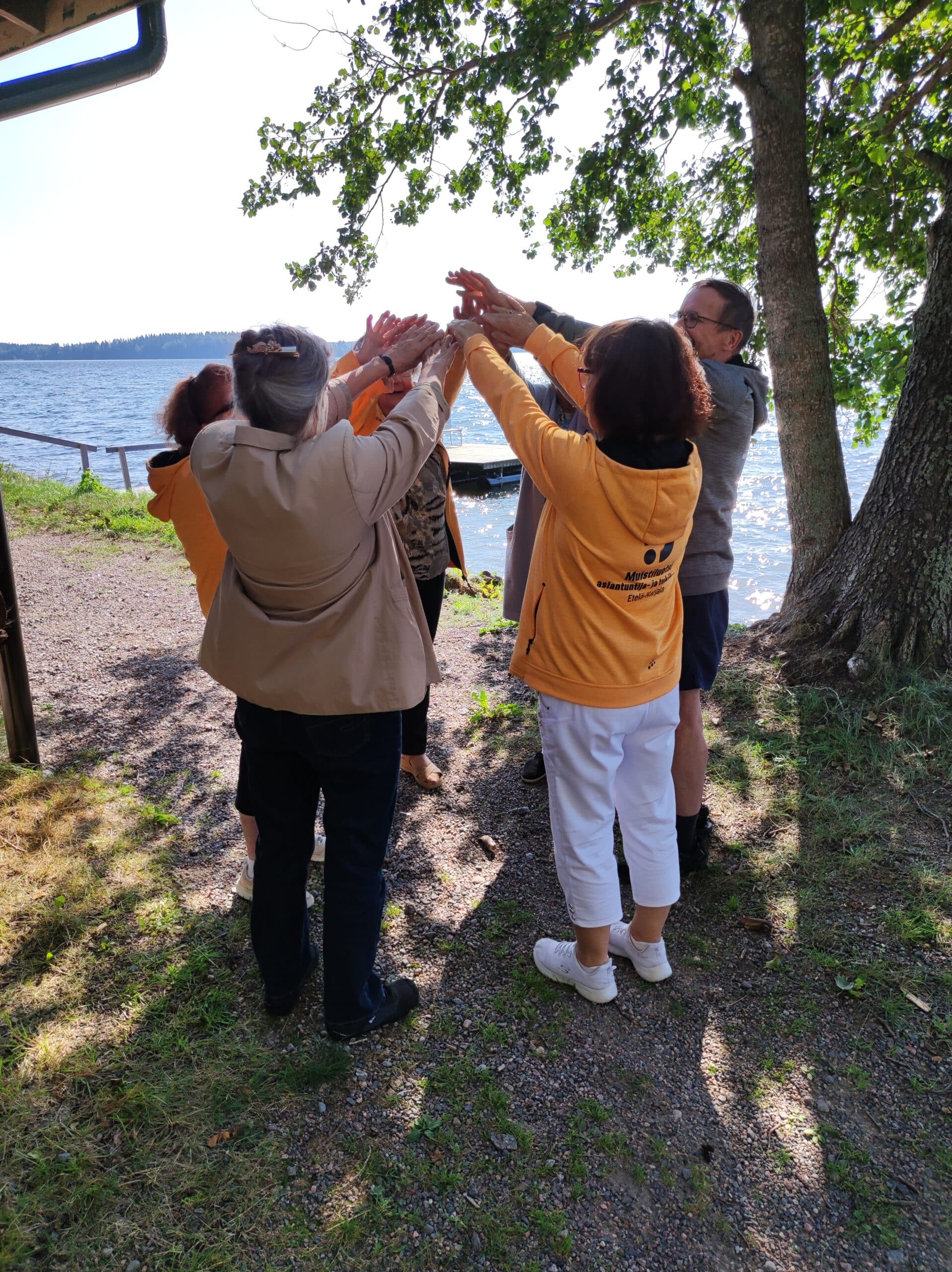 Ihmiset seisovat piirissä kädet kohotettuina järven rannalla