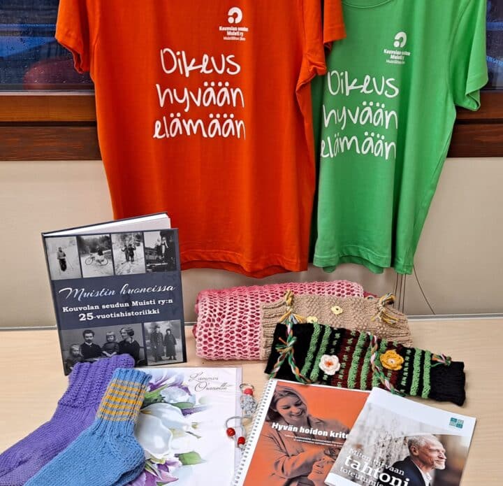 Kuvassa oranssi ja vihreä t-paita roikkumassa, pöydällä villasukkia, addressi, hypistelymuhveja ja kirjoja.