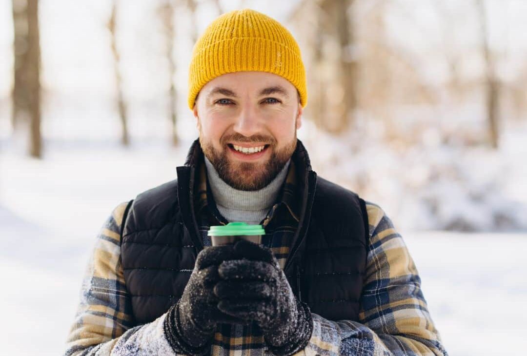 Kuvassa mies seisoo ulkona lumisessa maisemassa ja pitää molemmissa käsissään kahvikuppia. Hän katsoo kameraan ja hymyilee.