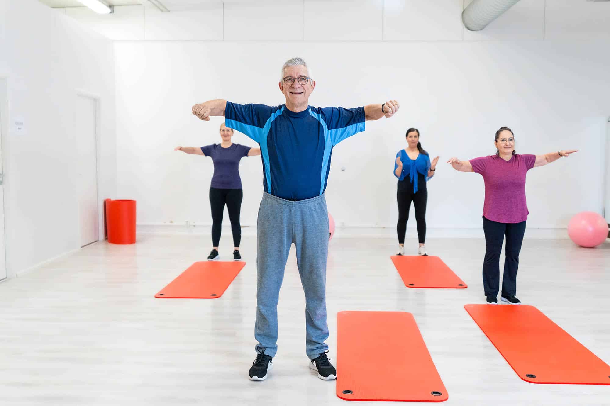 Liikunnan ryhmätunti. Sinipaitainen mies hymyilee etualalla. Taustalla näkyy muita liikkujia. Lattialla ion punaisia jumppamattoja.
