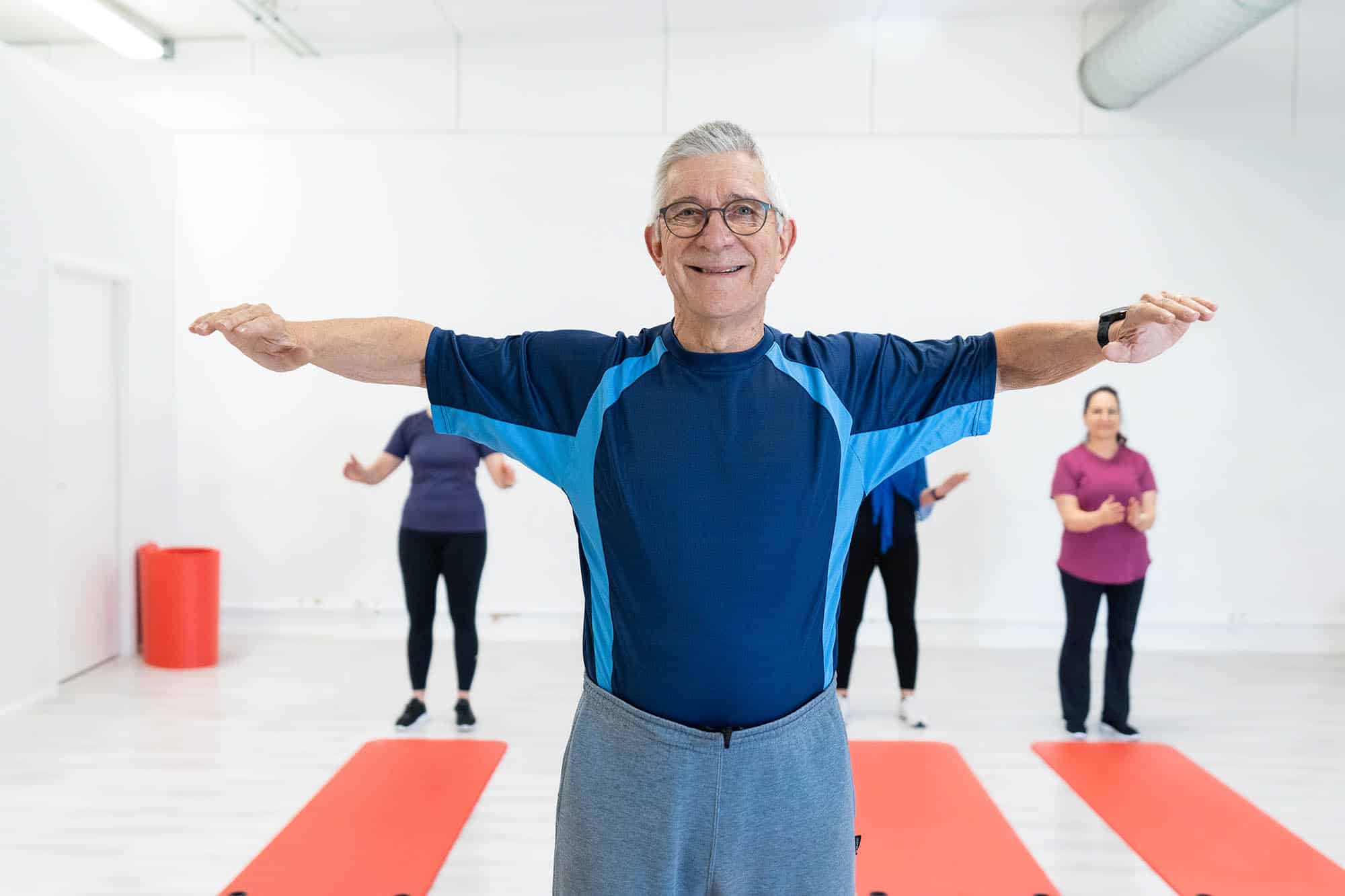 Liikuntasali, jossa on jumppamattoja ja jumppaajia. Etualalla sinpaitainen mies hymyilee ja katsoo kameraan, käsivarret nostettuina sivulle. Taustalla näkyy muita jumppaajia. Lattialla on jumppamattoja.