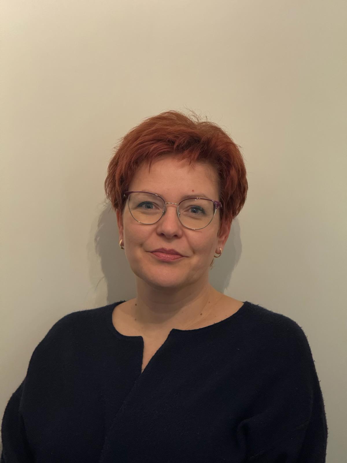 Kuvassa muistiohjaaja Camilla seisoo vaaleaa seinää vasten ja katsoo suoraan kameraan hymyillen.