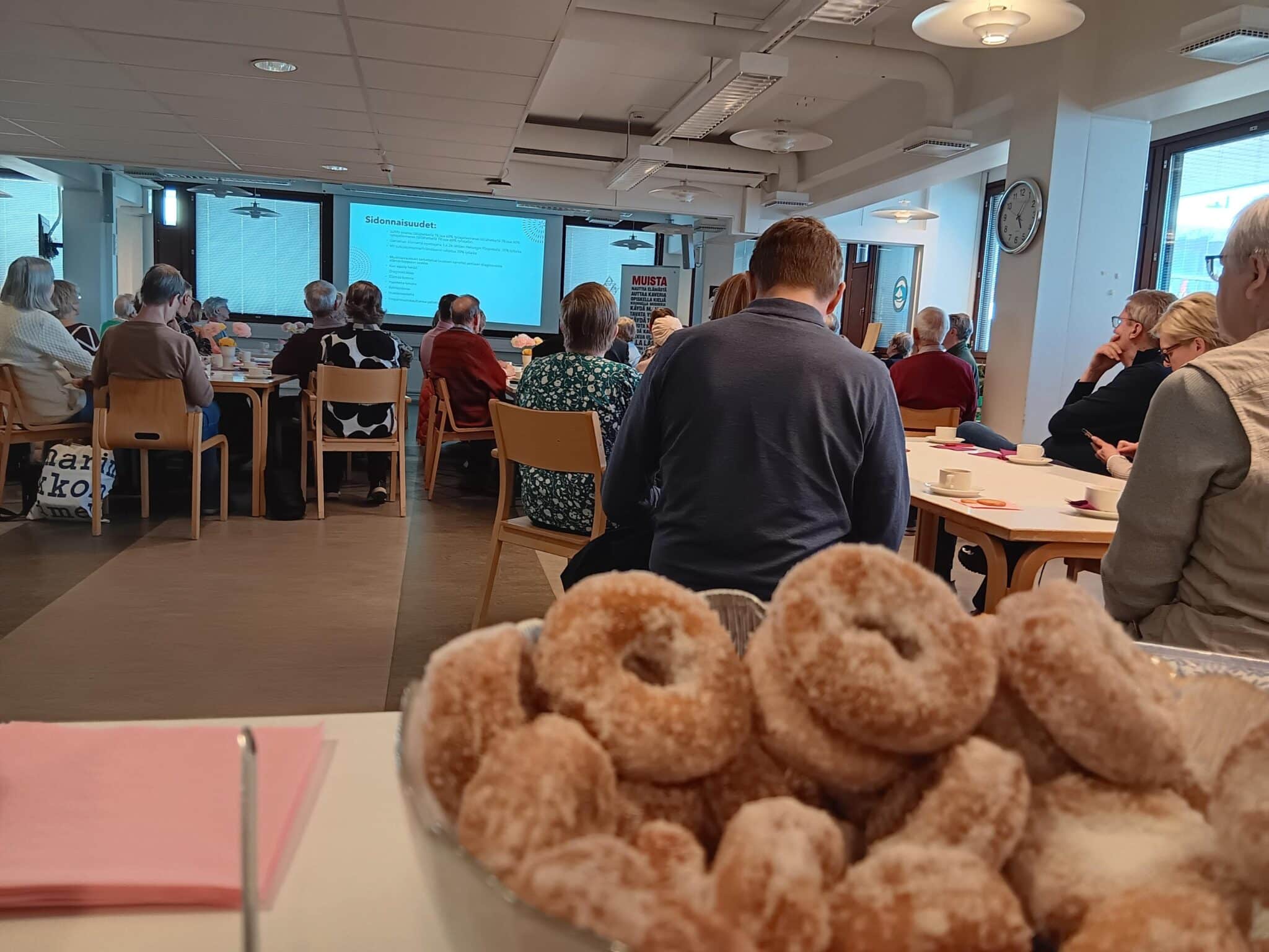 Kuvassa on Tapiolan palvelukeskuksen ruokasali, jossa on ihmisiä istumassa ja kuuntelemassa luentoa. Kuvan etualalla näkyy myös munkkeja, joita jaettiin kuuntelijoille vapun kunniaksi.