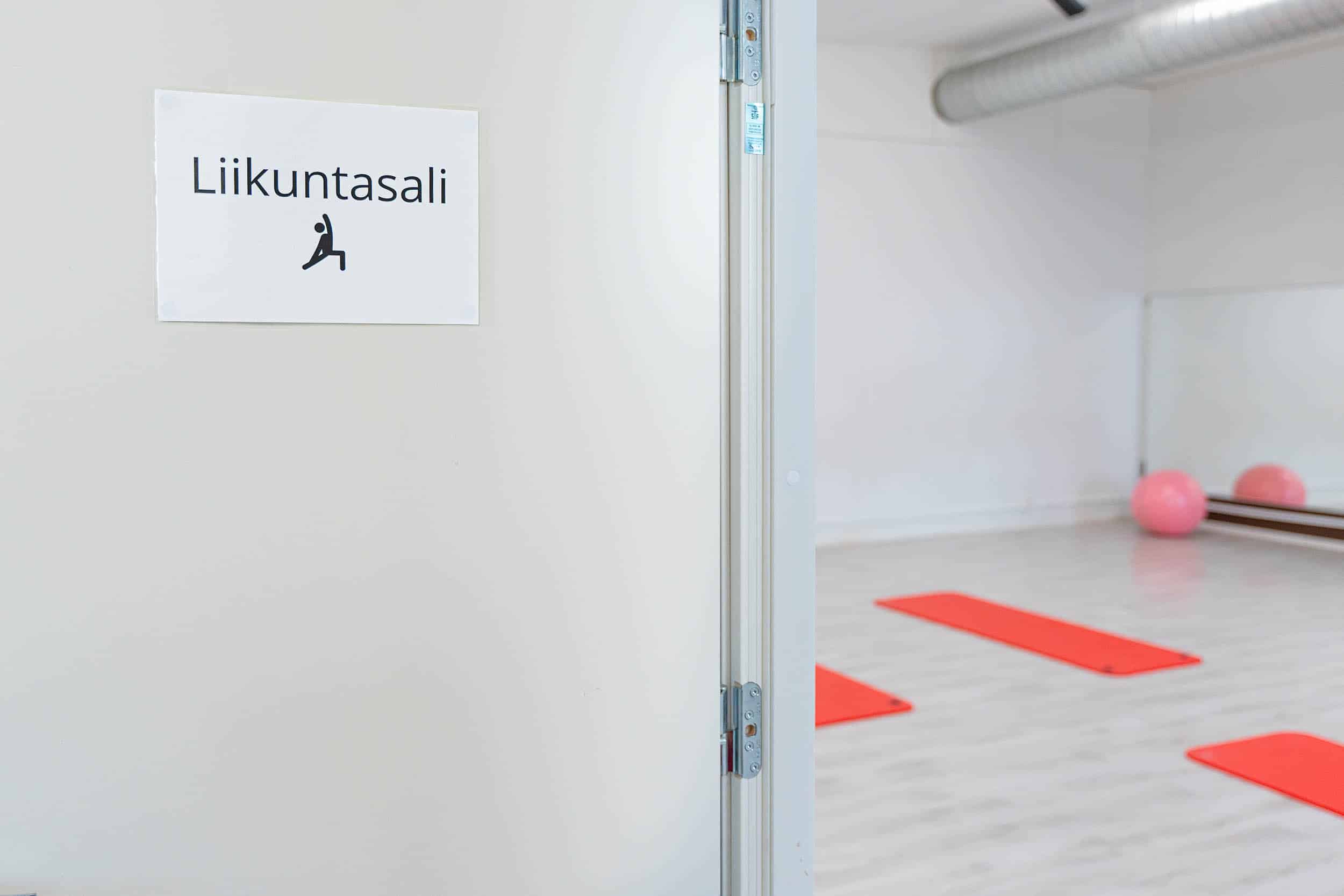 Näkymä ovelta liikuntasaliin, jossa näkyy urheiluvälineitä. Oveen on kiinnitetty opaste, jossa lukee: liikuntasali.